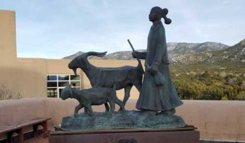 Bronze statue of a woman walking with a goat and a sheep, set against a mountainous backdrop.