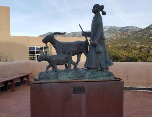 Bronze sculpture of a woman walking with a goat and a dog, set against a mountainous backdrop.