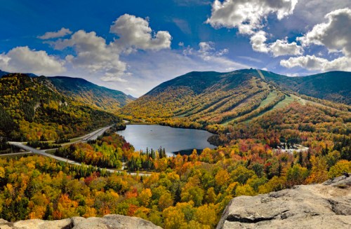 A panoramic view of colorful autumn foliage surrounding a serene lake, with mountains and a clear blue sky in the background.