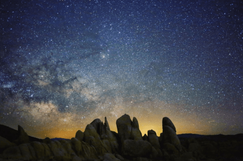 A starry night sky filled with stars and the Milky Way, above rocky formations and a distant horizon.