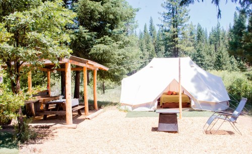 A cozy campsite featuring a white tent, wooden picnic area, and a chair surrounded by tall trees and greenery.