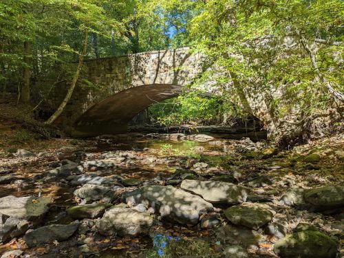 A stone bridge arches over a shallow stream, surrounded by lush green trees and rocky riverbed.