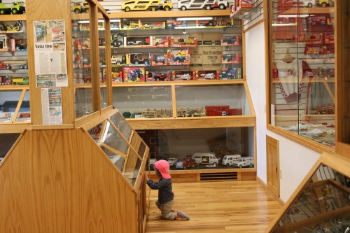 A child in a pink hat kneels by a display case in a toy museum filled with colorful toy cars and models.