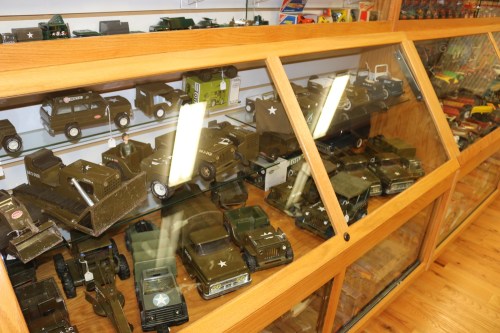 Display case filled with various military toy vehicles, including trucks and jeeps, arranged on wooden shelves.