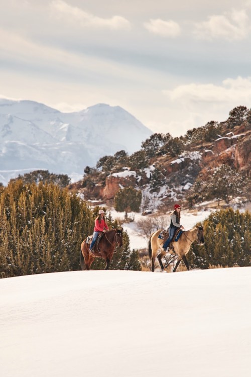 Two riders on horseback traverse a snowy landscape with mountains and trees in the background.