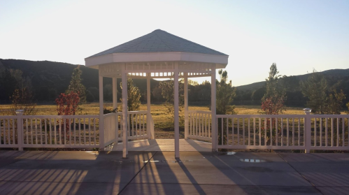 A white gazebo stands in a sunlit landscape, surrounded by trees and open fields under a clear sky.