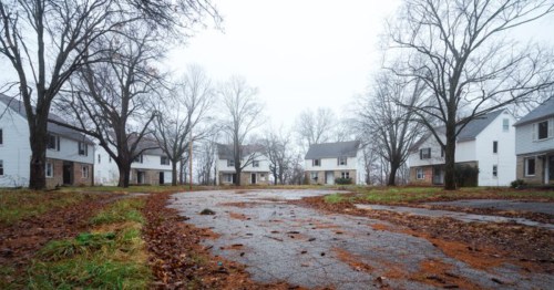 A foggy, deserted street lined with empty houses and bare trees, with fallen leaves scattered on the ground.