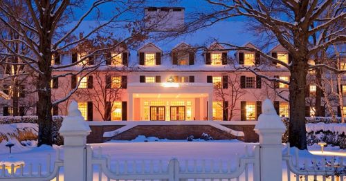 A grand pink hotel illuminated at dusk, surrounded by snow-covered trees and a white fence.