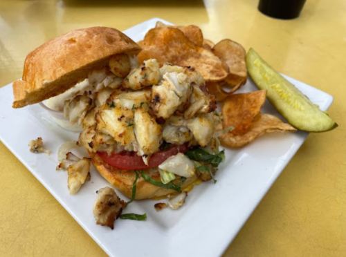A crab sandwich on a bun with lettuce and tomato, served with potato chips and a pickle on the side.
