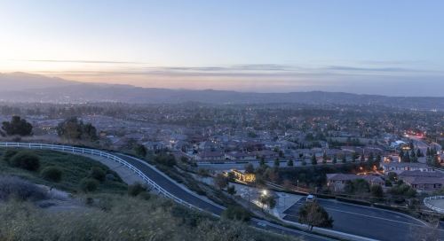 A panoramic view of a suburban landscape at dusk, with houses, winding roads, and distant mountains under a soft sky.