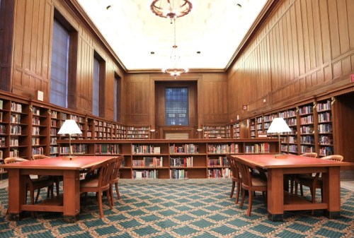 A spacious library with wooden walls, bookshelves, and tables, illuminated by lamps and chandeliers.