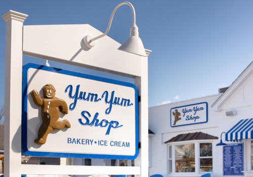 Sign for "Yum-Yum Shop" featuring a gingerbread man, with a bakery and ice cream theme in a sunny outdoor setting.