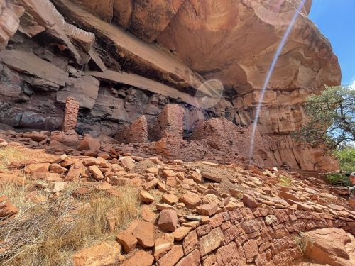 Ancient stone ruins nestled under a rocky overhang, surrounded by red earth and sparse vegetation.