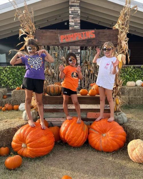 Three children with face paint stand on large pumpkins, smiling and posing at a pumpkin patch.