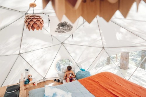A cozy geodesic dome interior with a child sitting on the floor, surrounded by soft furnishings and natural light.