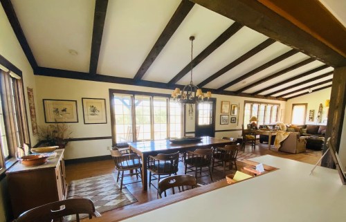 A spacious dining area with a long table, wooden beams, and large windows, leading to a cozy living room.