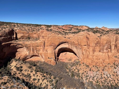 A stunning view of a large sandstone arch in a desert landscape, surrounded by red rock formations and clear blue skies.