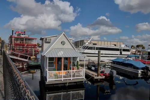 A small houseboat with a porch is docked beside larger boats in a marina under a partly cloudy sky.