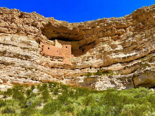 Ancient cliff dwellings nestled in rocky cliffs, surrounded by green vegetation under a clear blue sky.