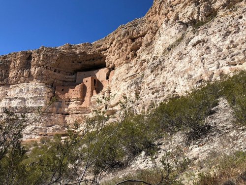 Ancient cliff dwellings nestled in a rocky landscape under a clear blue sky, surrounded by sparse vegetation.