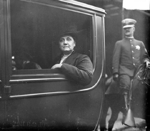 A historical black-and-white photo of a woman in a car, with a man in a uniform on horseback in the background.