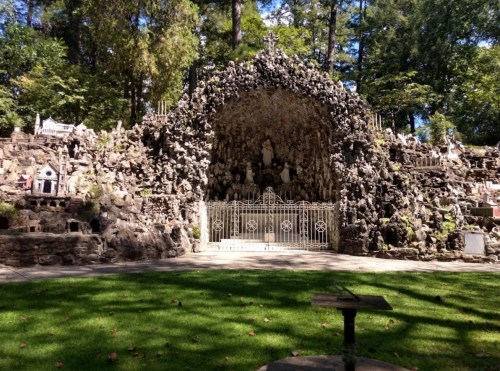 A stone grotto adorned with statues and small structures, surrounded by greenery and trees.