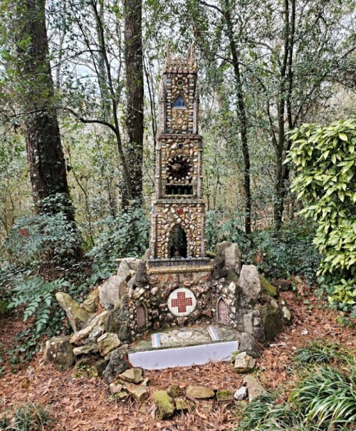 A decorative stone structure surrounded by trees and greenery, featuring intricate designs and a cross at its base.