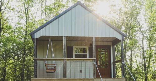 A small, modern cabin with a porch swing, surrounded by trees and sunlight filtering through the leaves.