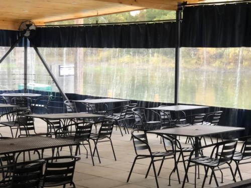 Empty outdoor dining area with black tables and chairs, overlooking a calm body of water through sheer curtains.