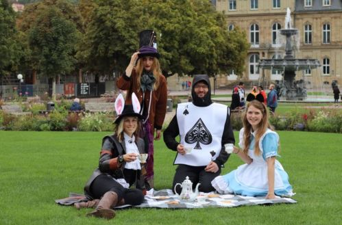 A group dressed as Alice in Wonderland characters enjoys a tea party in a park, surrounded by greenery and a fountain.