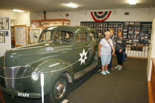 Three women stand beside a vintage green military vehicle in a museum, surrounded by historical displays.