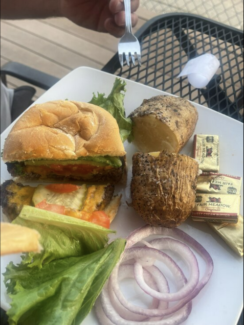 A plate with a burger, lettuce, tomato, onion slices, and two baked potatoes, with a fork resting on the side.