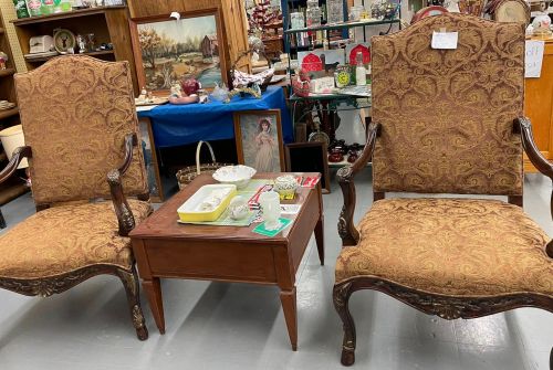 Two ornate chairs with patterned upholstery flanking a small wooden table, surrounded by various antiques and decor.