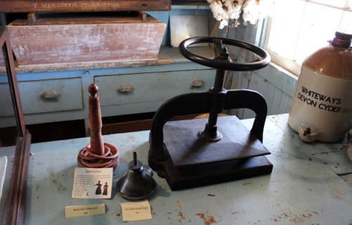 A vintage press and two small objects on a wooden table, with informational cards beside them.