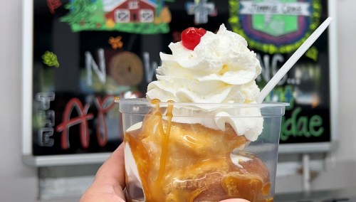 A hand holding a cup of ice cream topped with whipped cream and a cherry, with a colorful chalkboard in the background.