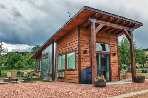 A modern wooden cabin with large windows, surrounded by gravel and greenery, under a cloudy sky.