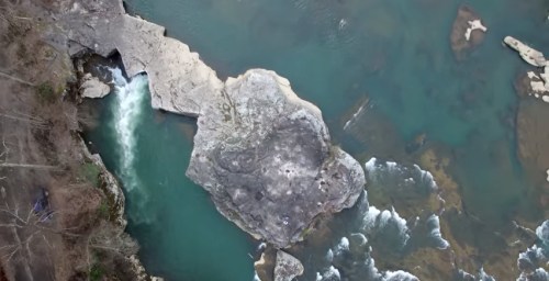 Aerial view of a rocky riverbank with flowing water and a large stone formation in the center.