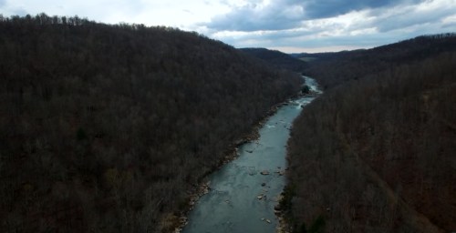 Aerial view of a winding river surrounded by bare trees and rolling hills under a cloudy sky.