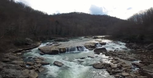 A serene river flows over rocky formations, surrounded by trees and hills under a cloudy sky.