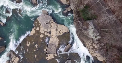 Aerial view of a river with rocky formations and flowing water, surrounded by trees and a rocky bank.