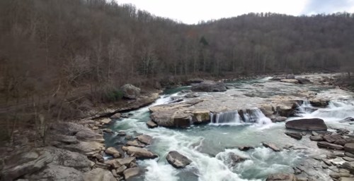 A serene river flows over rocky terrain, surrounded by bare trees and a hilly landscape under a cloudy sky.