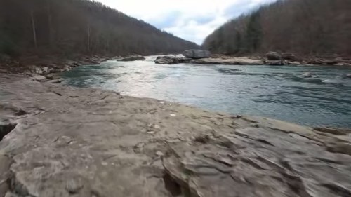 A rocky riverbank with calm water flowing through a scenic landscape of trees and hills in the background.