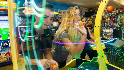 Two young people play an arcade game, focused and smiling, with colorful lights reflecting around them.