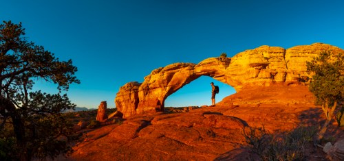 A person stands beneath a natural rock arch at sunset, surrounded by red rock formations and clear blue skies.