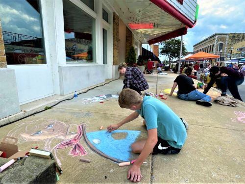 Children and teens create colorful chalk art on a sidewalk outside a building during a community event.