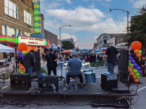 A band performs on a stage in a lively street festival, with colorful balloons and a crowd enjoying the event.