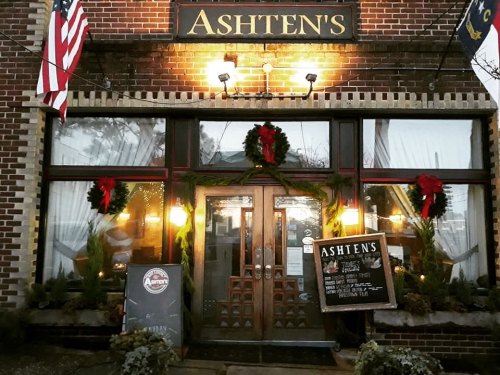 A cozy restaurant entrance decorated with wreaths and lights, featuring a sign that reads "Ashten's." Flags are displayed.