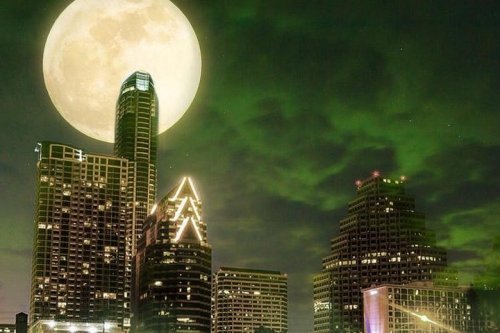 A large full moon illuminates a city skyline at night, with greenish clouds adding a dramatic effect.