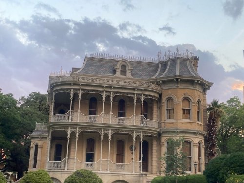 Victorian-style mansion with ornate balconies, surrounded by greenery under a cloudy sky.