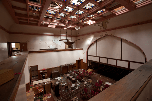 Interior of a dilapidated room with a damaged ceiling, stained glass, scattered furniture, and debris on the floor.
